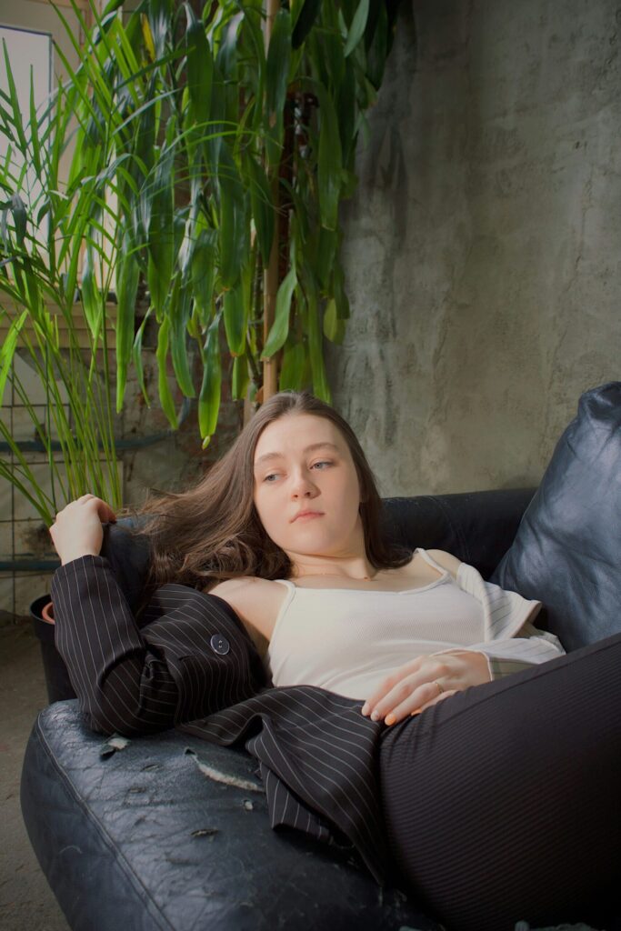 Woman with brown hair, light skin, and a sullen face, reclining on a couch with a green plant behind her.