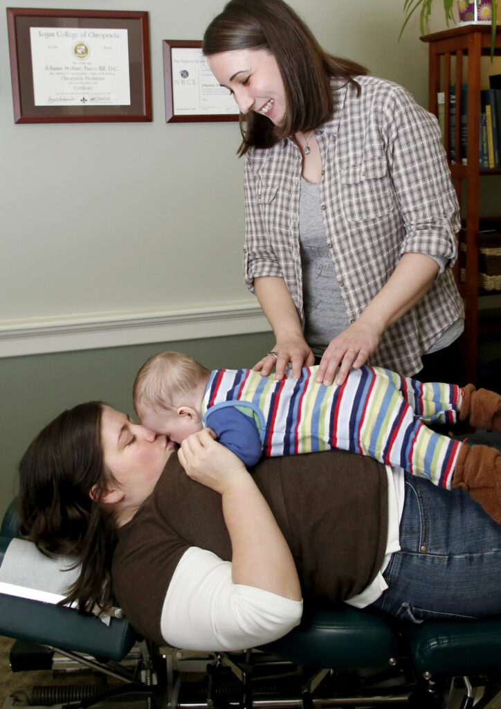 Mom laying on her back on a treatment table with her baby on her belly. She is lifting her head and kissing her baby while a smiling female doctor stands over them touching the baby's back.