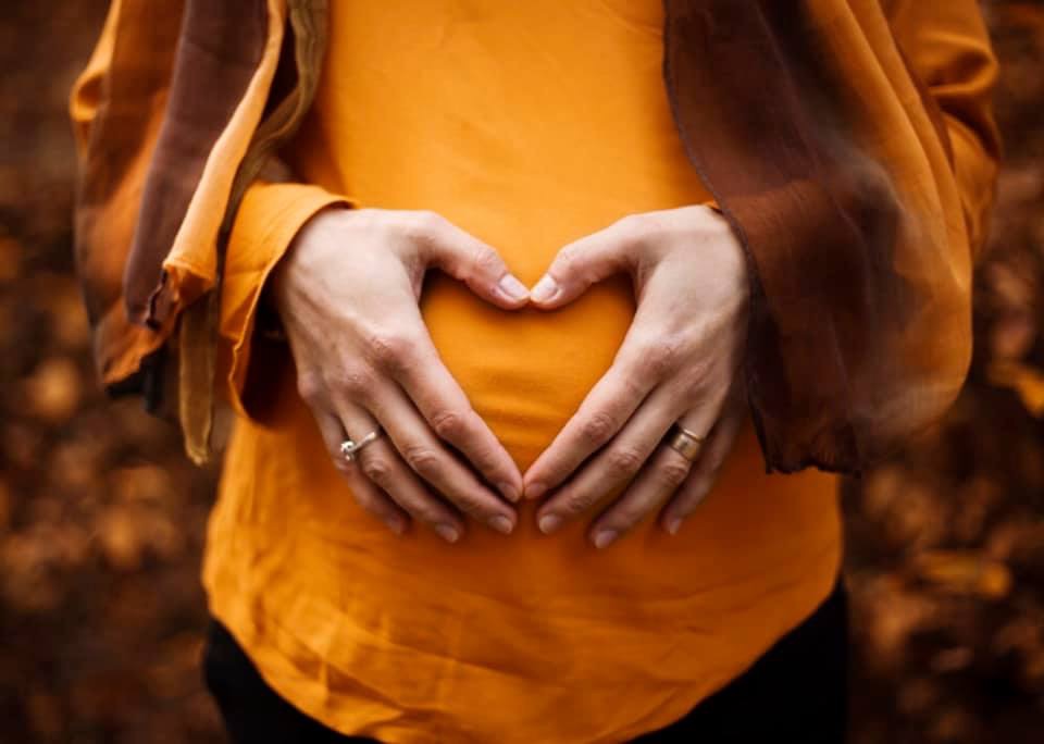 Image of a pregnant belly with the person wearing all orange and holding their hands in a heart shape on their belly