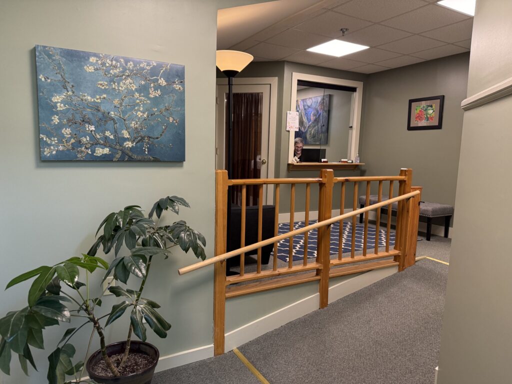 Image of a waiting room from the view down a ramp looking up into the room. There is a large green plant on the left below a painting of cherry blossoms, a bench, rug, and couch are visible behind the wooden railing, as well as a painting and the front desk window with a smiling woman behind the window